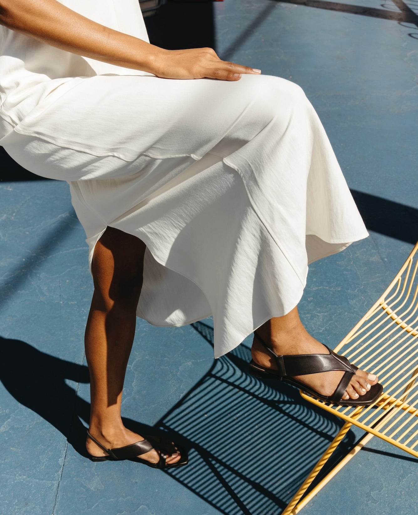 Lower half of a woman in a white dress with her right leg propped on a yellow wire chair and feet wearing the Finch Sandal in Black.