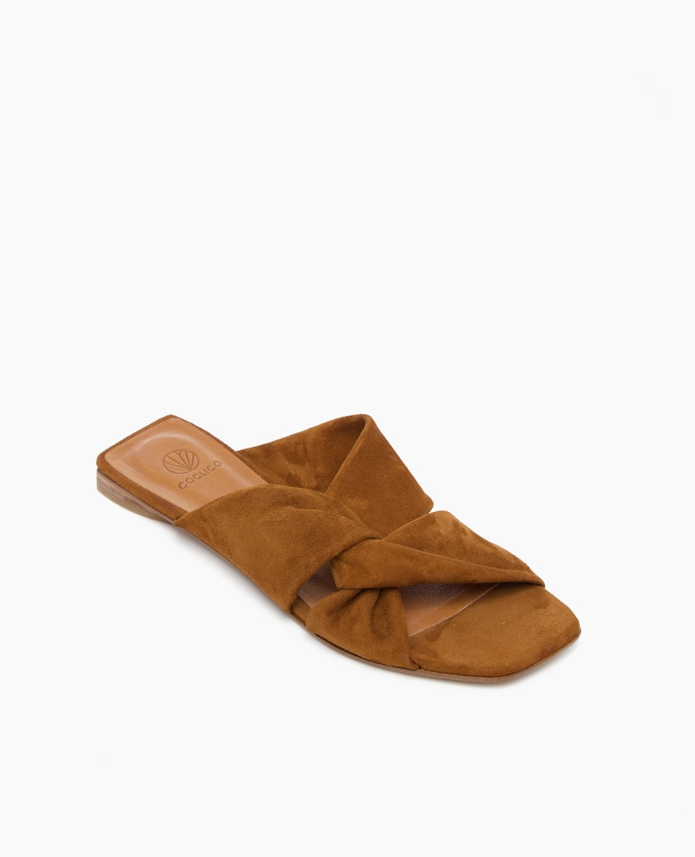 A brown suede slide sandal features a twisted strap design, a flat sole, and a square open toe, with a smooth leather insole.