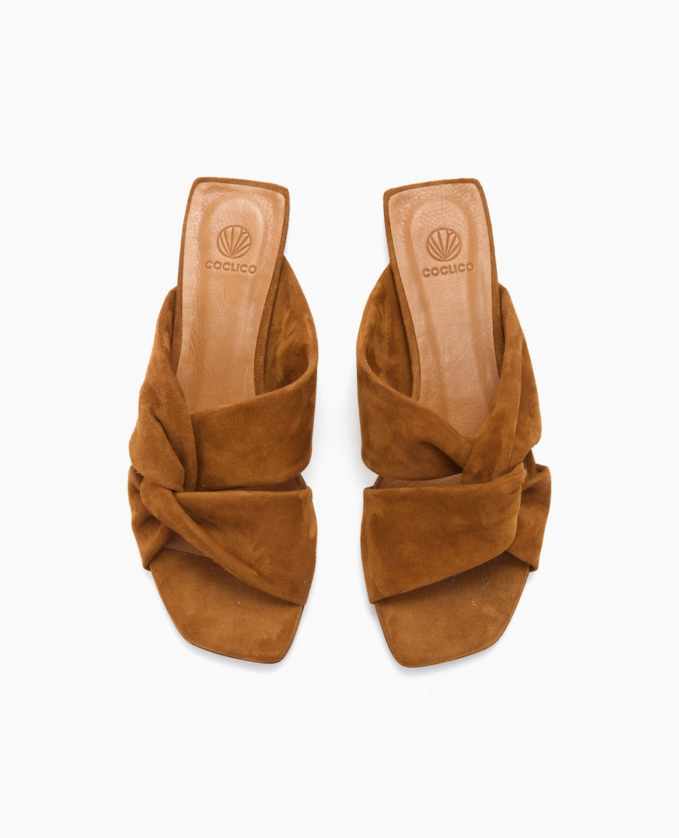 Brown suede sandals with a twisted knot design, featuring a square open toe and a flat sole, displaying a soft, textured finish and a branded insole.