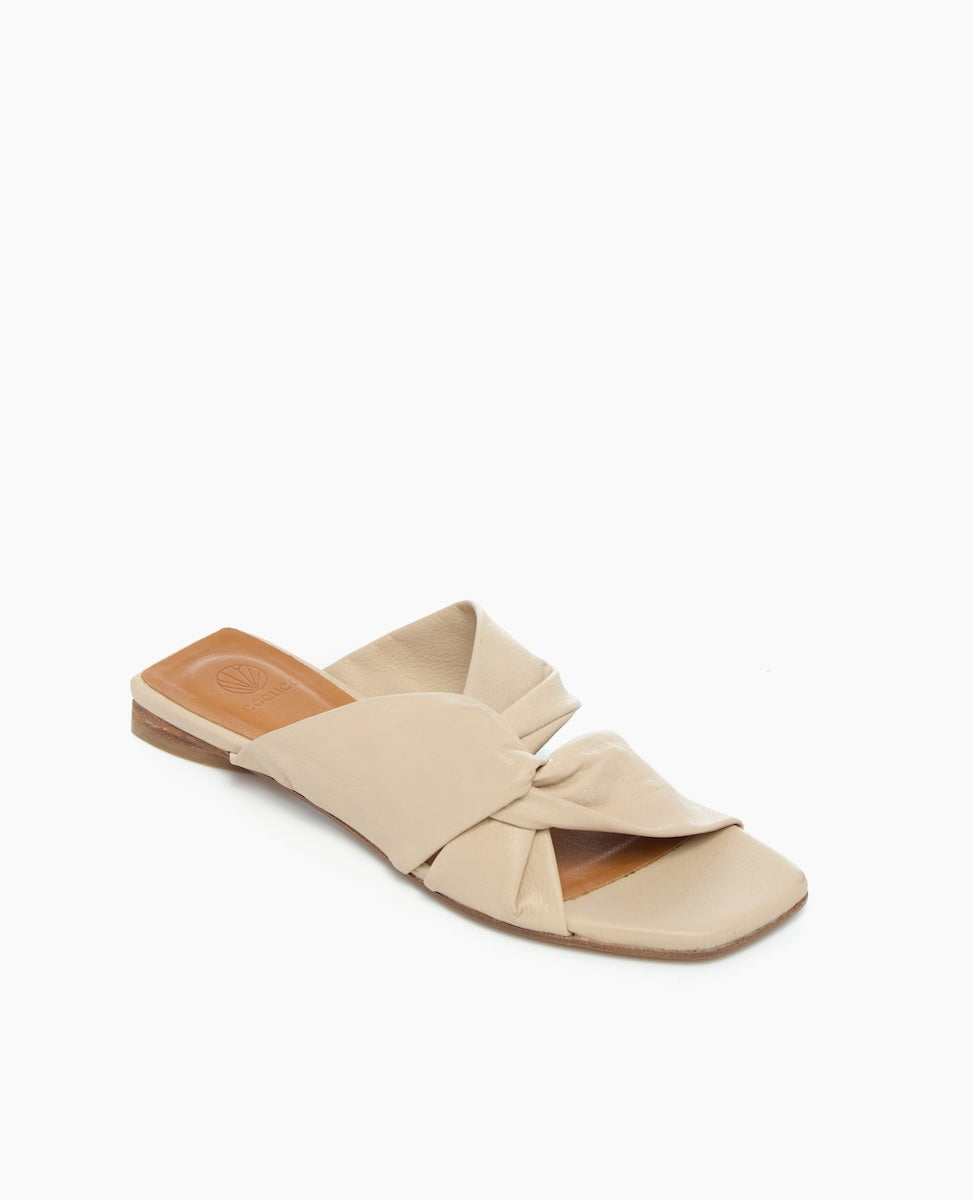 A beige, flat slide sandal features a soft, twisted leather upper with an open toe design and a smooth, tan insole.