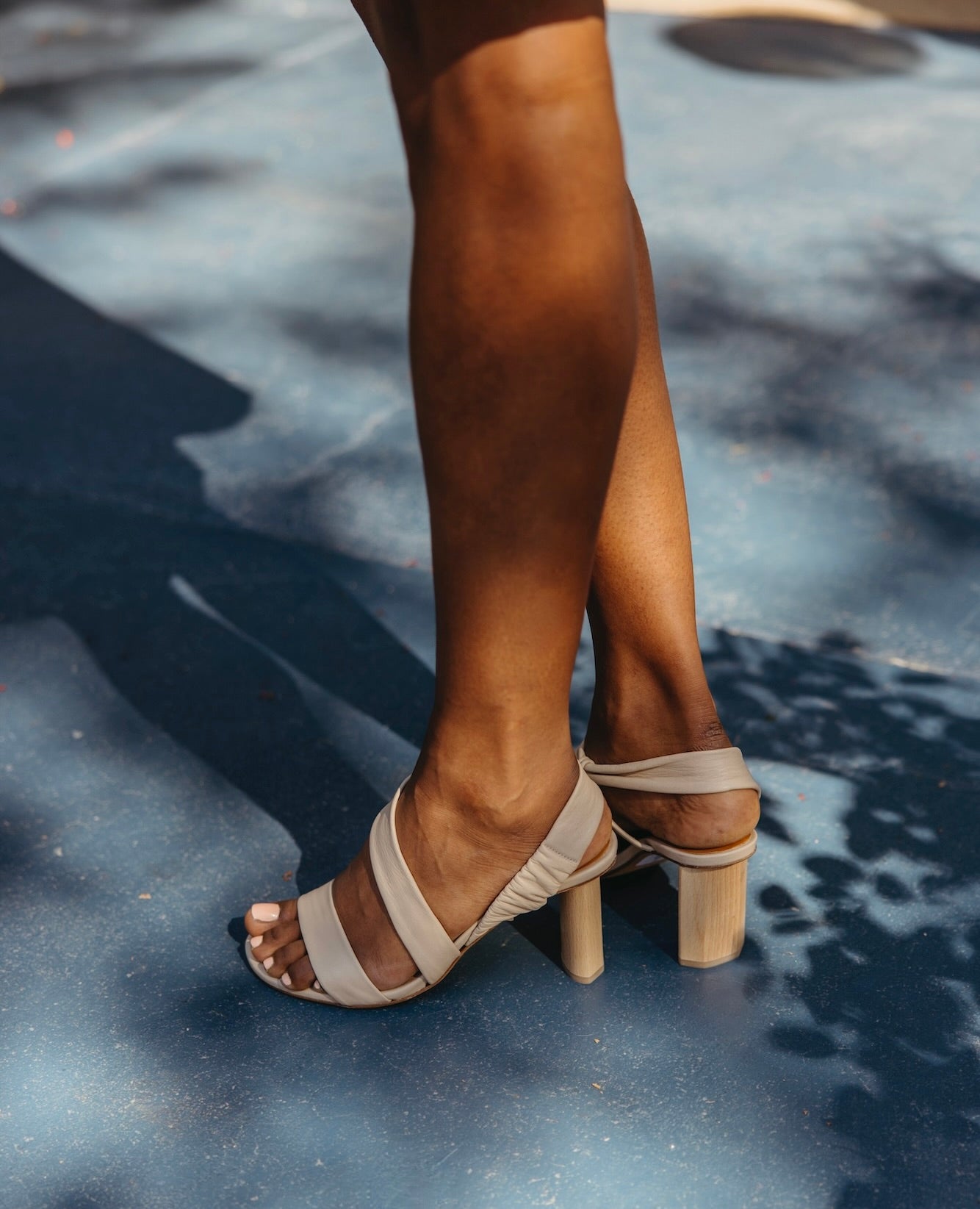 Legs of a woman standing on a blue concrete floor wearing the Gabby Heel in Latte Macchiato.