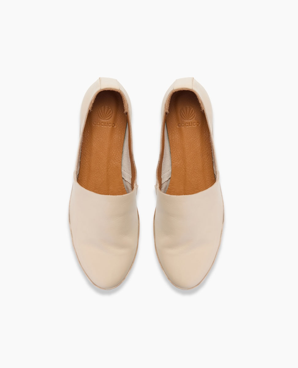 A pair of cream-colored slip-on shoes with a smooth, rounded toe, tan interior lining, and a subtle logo embossed on the insole.