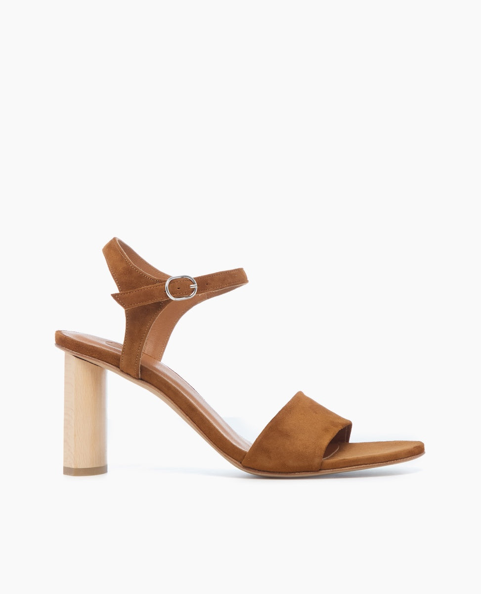 A brown suede sandal with an open toe, featuring a wide front strap, an adjustable ankle strap with a silver buckle, and a distinctive cylindrical wooden block heel.