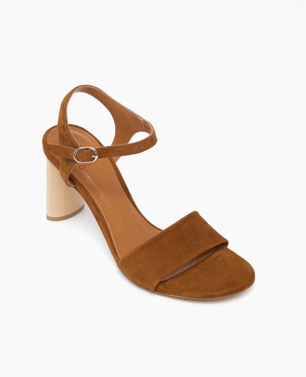 A brown suede sandal with an open toe, featuring a wide front strap, an adjustable ankle strap with a silver buckle, and a cylindrical wooden block heel.
