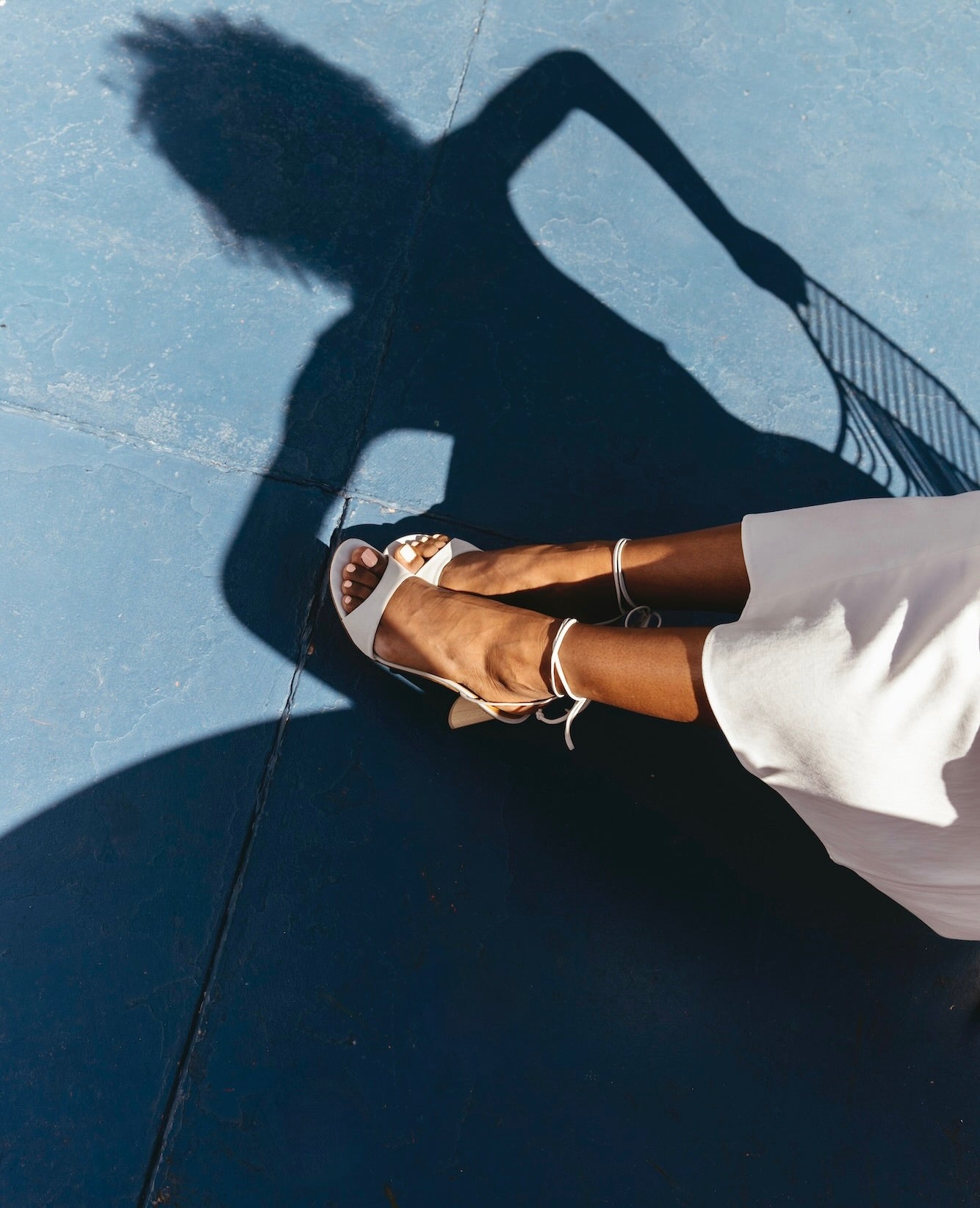 Legs and feet stretched out wearing the Gilda in Griege Nubuck with half of a white skirt pictured against a blue concrete floor.