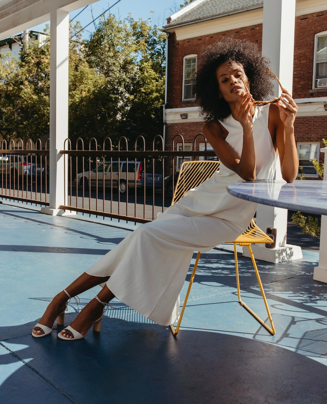 Woman in a white dress and the Gilda in Griege Nubuck holding sunglasses and sitting at a table.