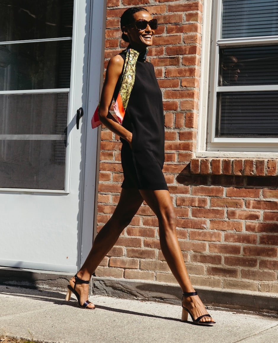 Woman wearing a black shift dress with the Gina Heel in Black Pearl Patent while walking outside.