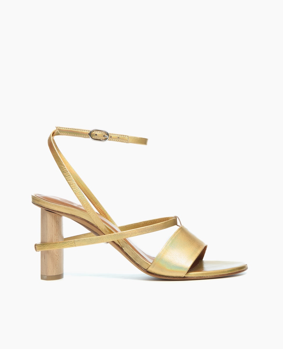A sleek, gold sandal featuring a minimalist design with thin straps, an adjustable ankle buckle, and a unique cylindrical wooden heel.