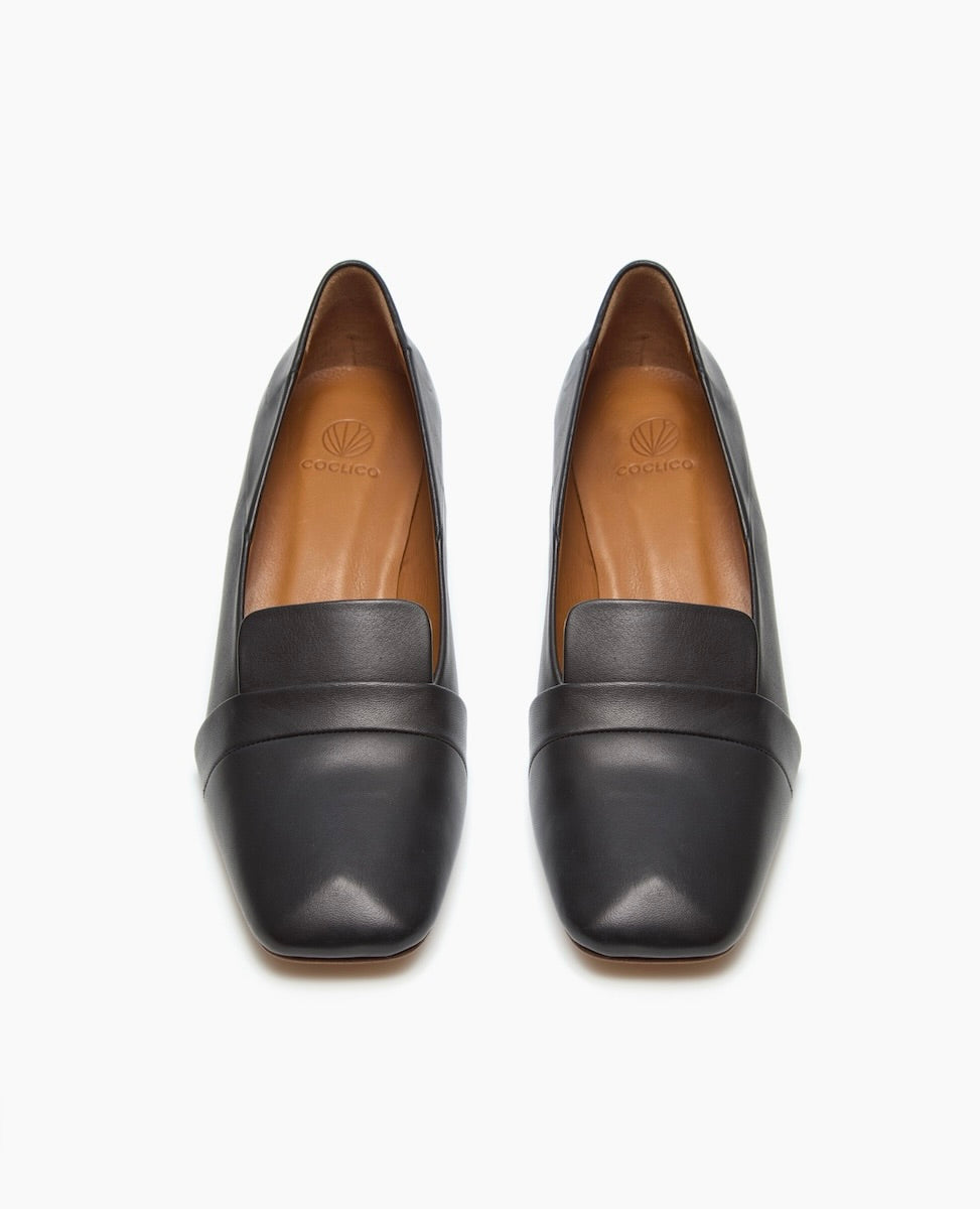 A pair of sleek, black leather loafers with a smooth texture, square toes, and a minimalist design featuring a subtle, raised band across the front and tan insoles with a logo imprint.