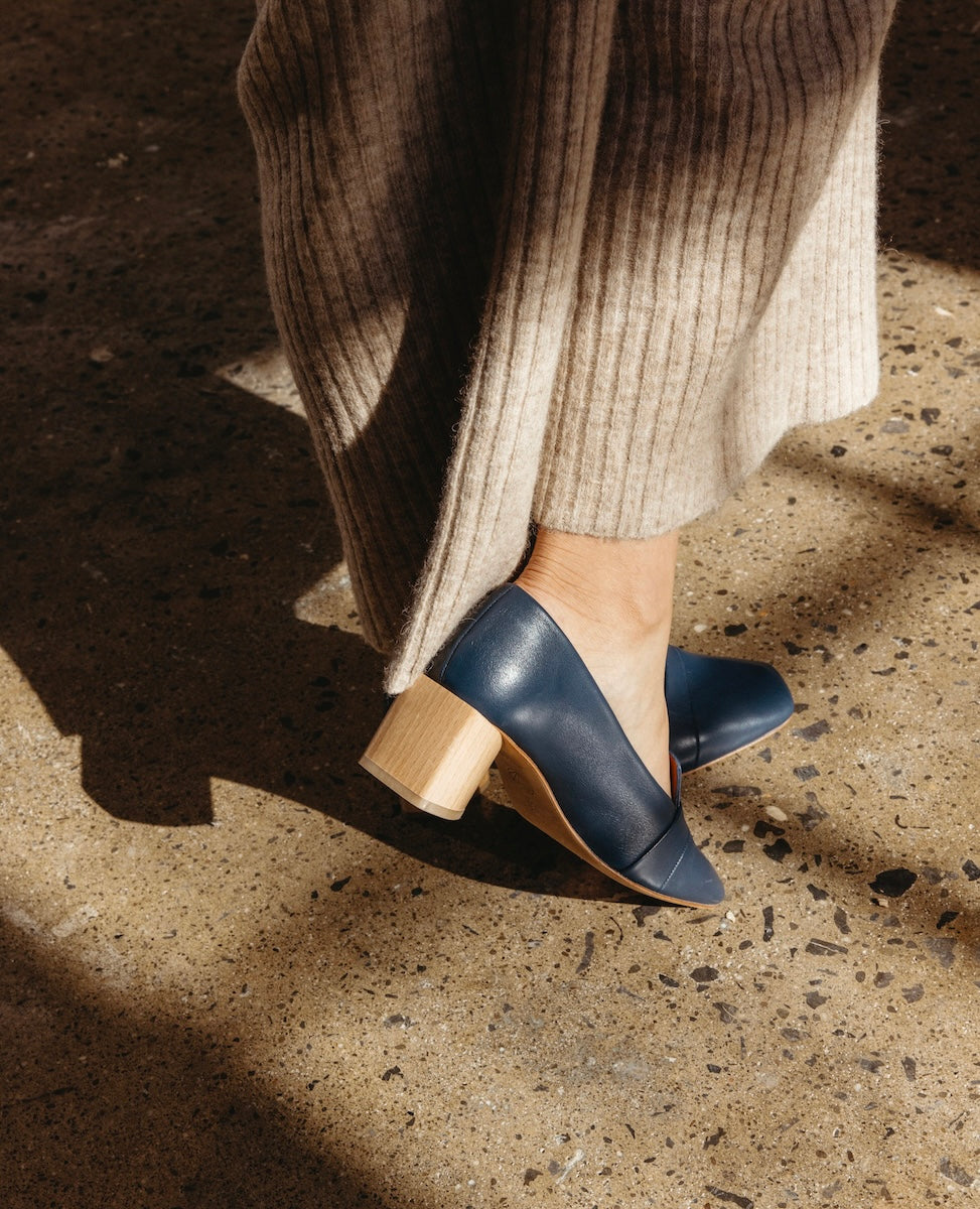 A pair of navy blue leather shoes with a rounded toe and a light wooden block heel, worn with beige ribbed pants on a textured concrete surface.
