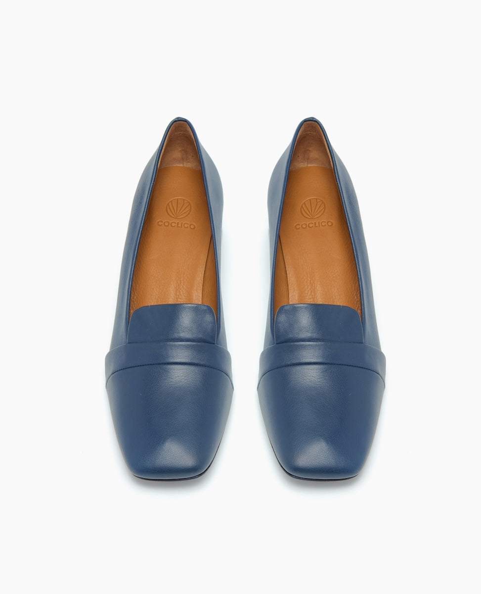 A pair of sleek, blue leather loafers with a smooth texture, almond-shaped toes, and tan insoles featuring a subtle logo imprint.