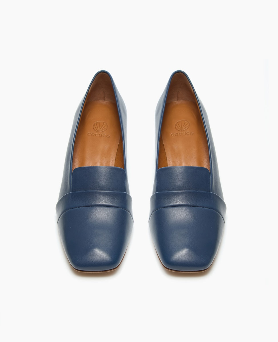 A pair of sleek, blue leather loafers with a square toe design, featuring a smooth texture and a subtle seam across the upper, lined with tan leather interiors.