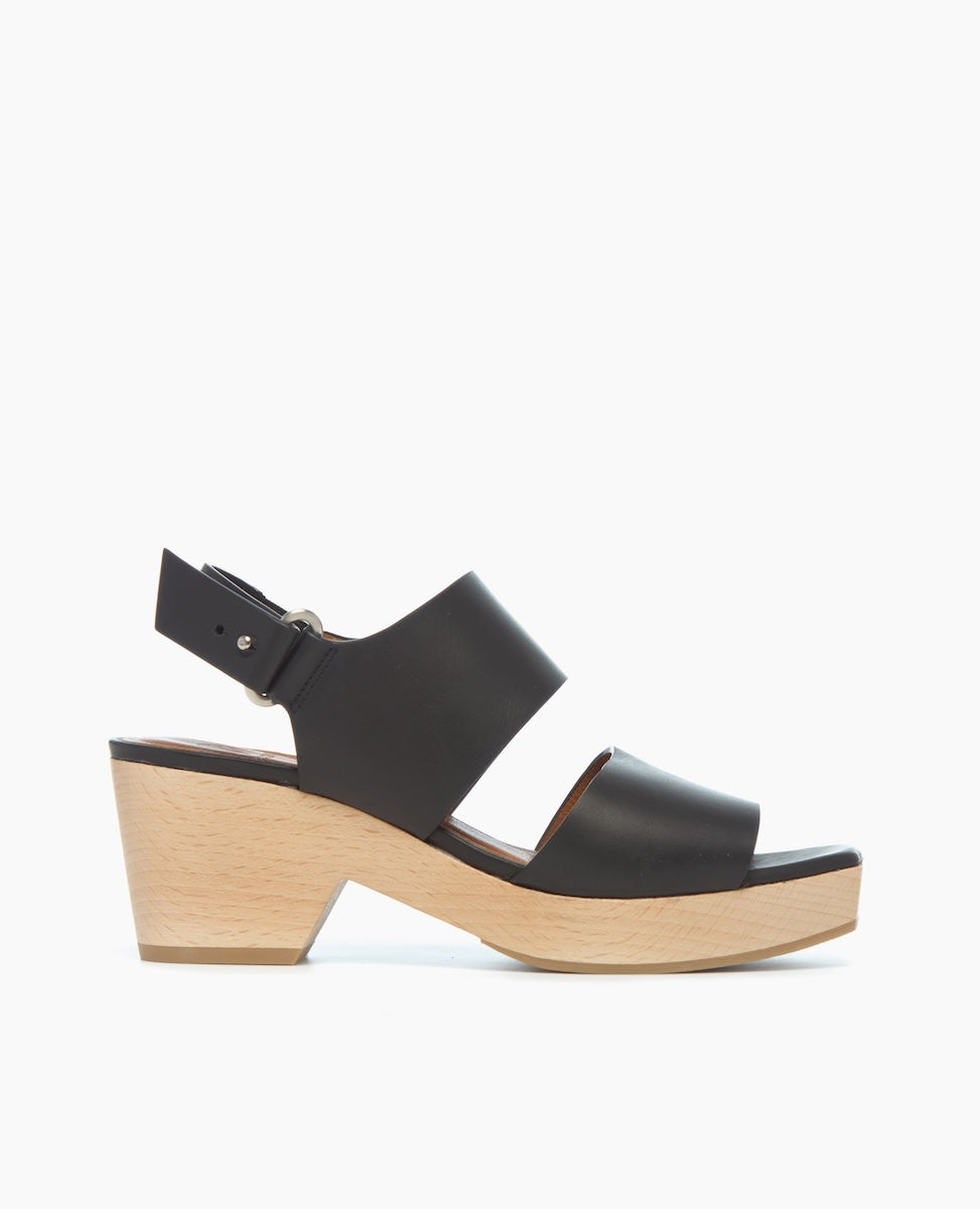 A black leather sandal with a wide strap design, featuring a chunky light wood platform heel and an adjustable ankle strap with a silver buckle.