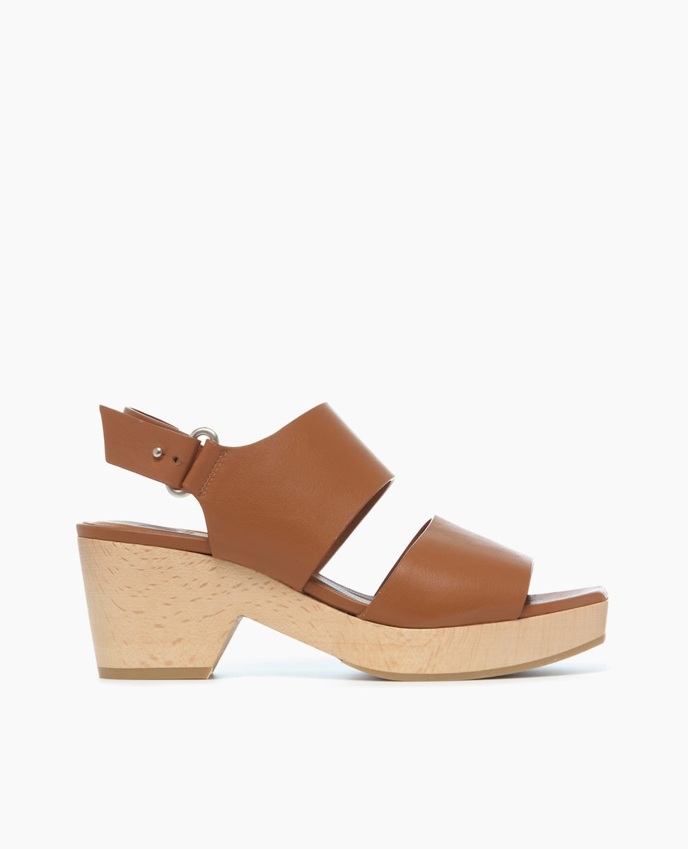 A brown leather sandal with a wide strap design, featuring a wooden wedge heel and an adjustable ankle strap with a silver buckle.