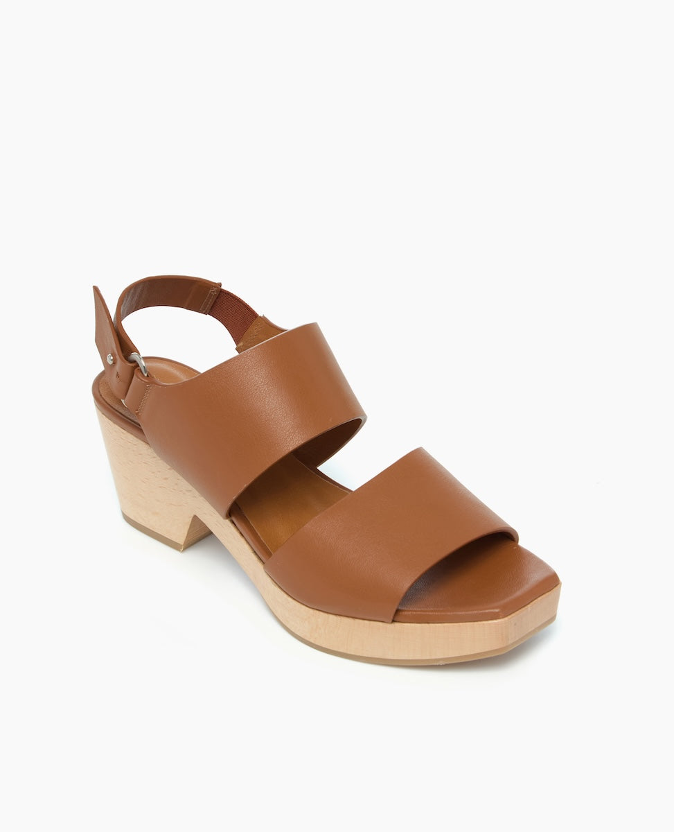 A brown leather sandal with a chunky wooden heel, featuring a wide front strap, an adjustable ankle strap with a buckle, and a smooth, matte finish.