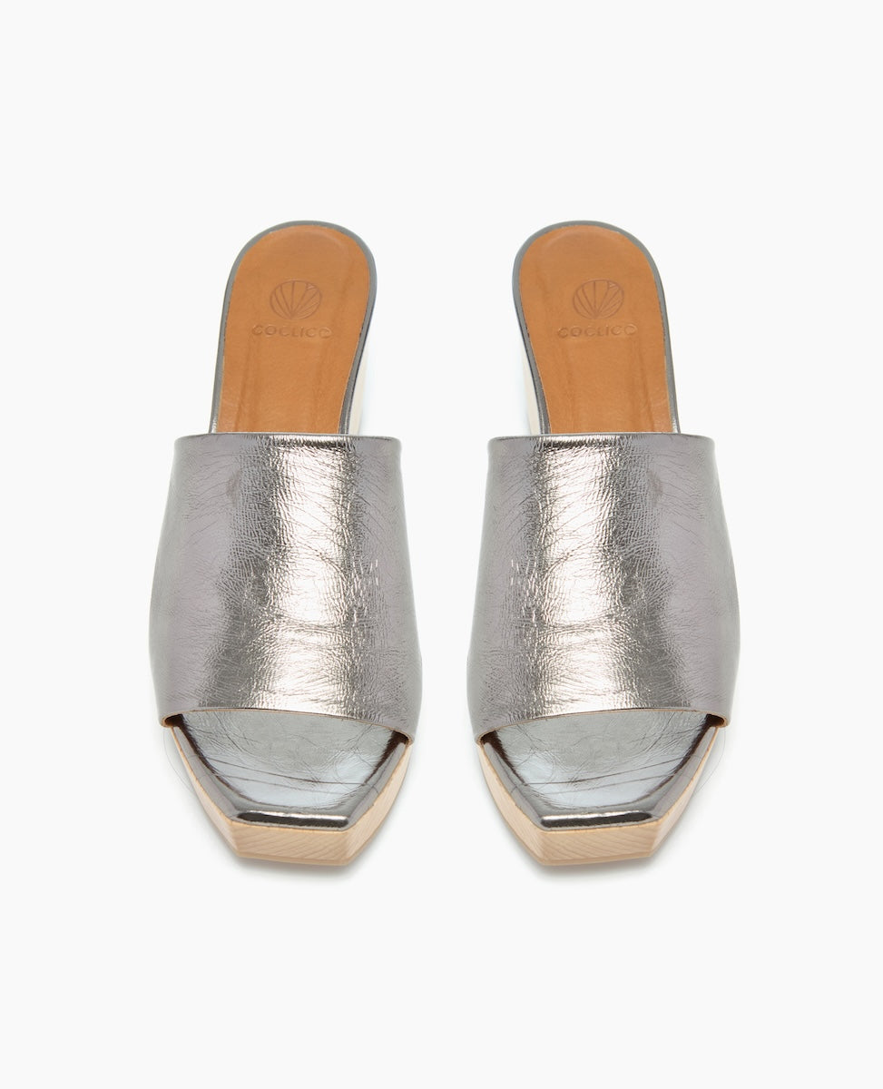Silver metallic open-toe mules with a smooth, shiny texture, featuring a tan insole and a low, light-colored wooden platform heel.