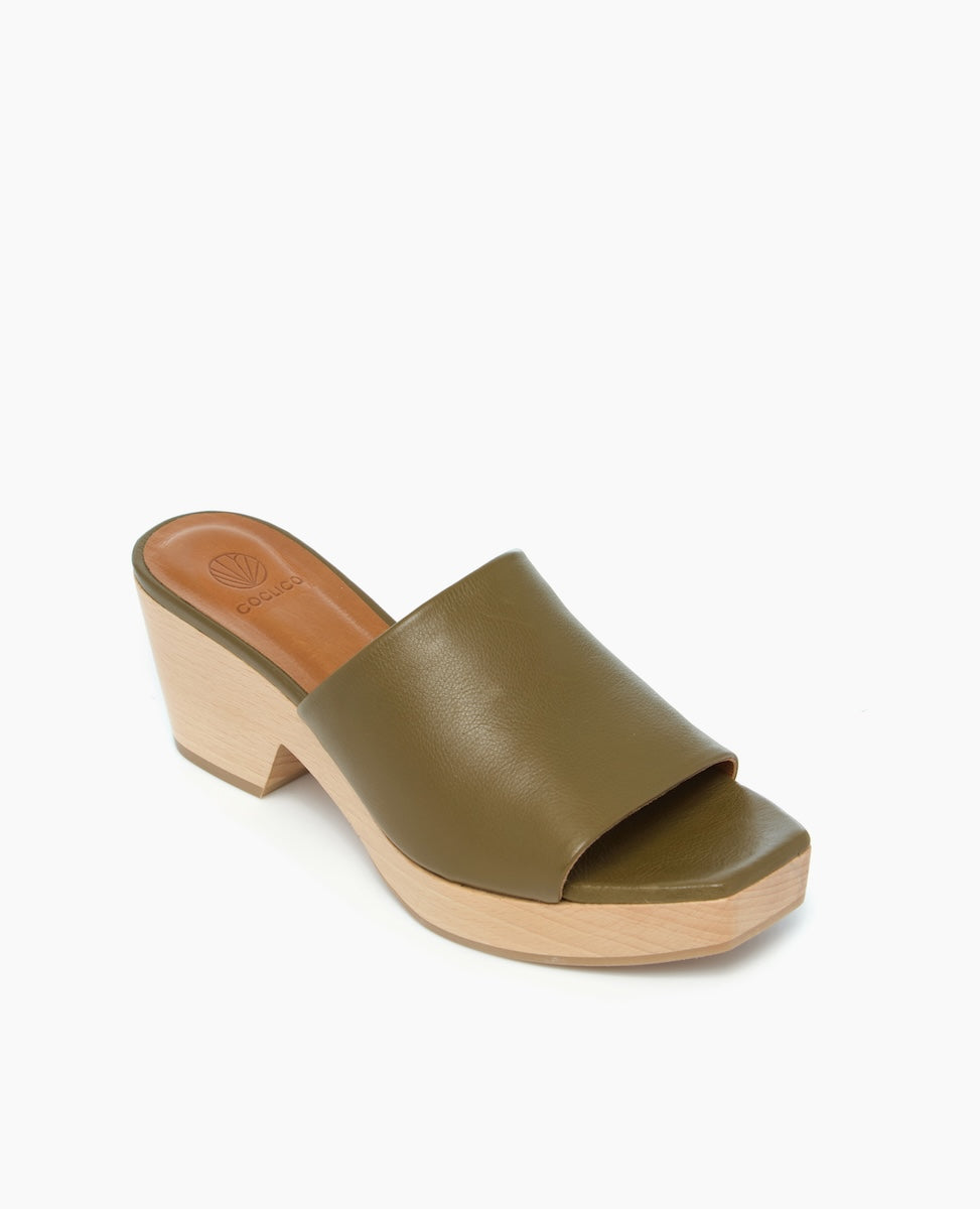 A stylish olive green leather mule with an open toe design, featuring a smooth texture, a wooden wedge heel, and a minimalist aesthetic.