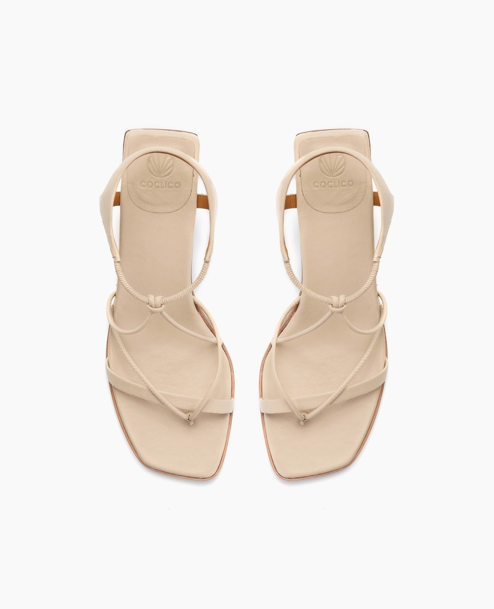 Beige sandals with a minimalist design feature thin straps, a square toe, and a flat sole, crafted from smooth leather with a subtle embossed logo on the insole.