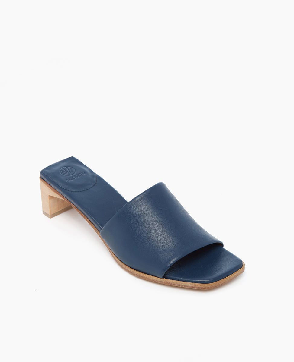 A sleek, navy blue leather slide sandal with a wide strap, open toe, square toe shape, and a low, light wooden block heel.