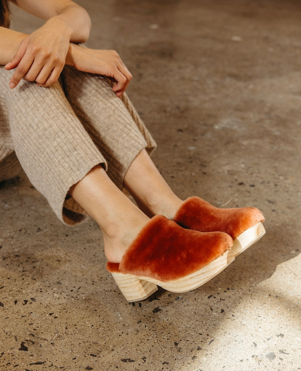 A person wearing beige pants is seated on a concrete floor, showcasing a pair of stylish, Autumn Leaf Shearling with wooden soles.