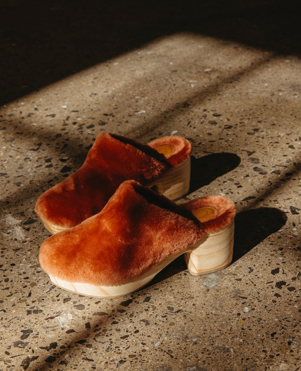 A pair of Autumn Leaf Shearling clogs with wooden soles is placed on a speckled concrete floor, illuminated by sunlight.