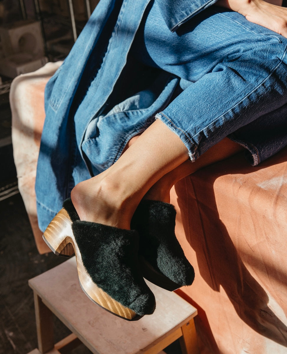 A pair of Hunter Green Shearling slip-on clogs with wooden soles and a slightly elevated heel, worn by a person in blue jeans and a denim jacket, resting on a wooden stool.
