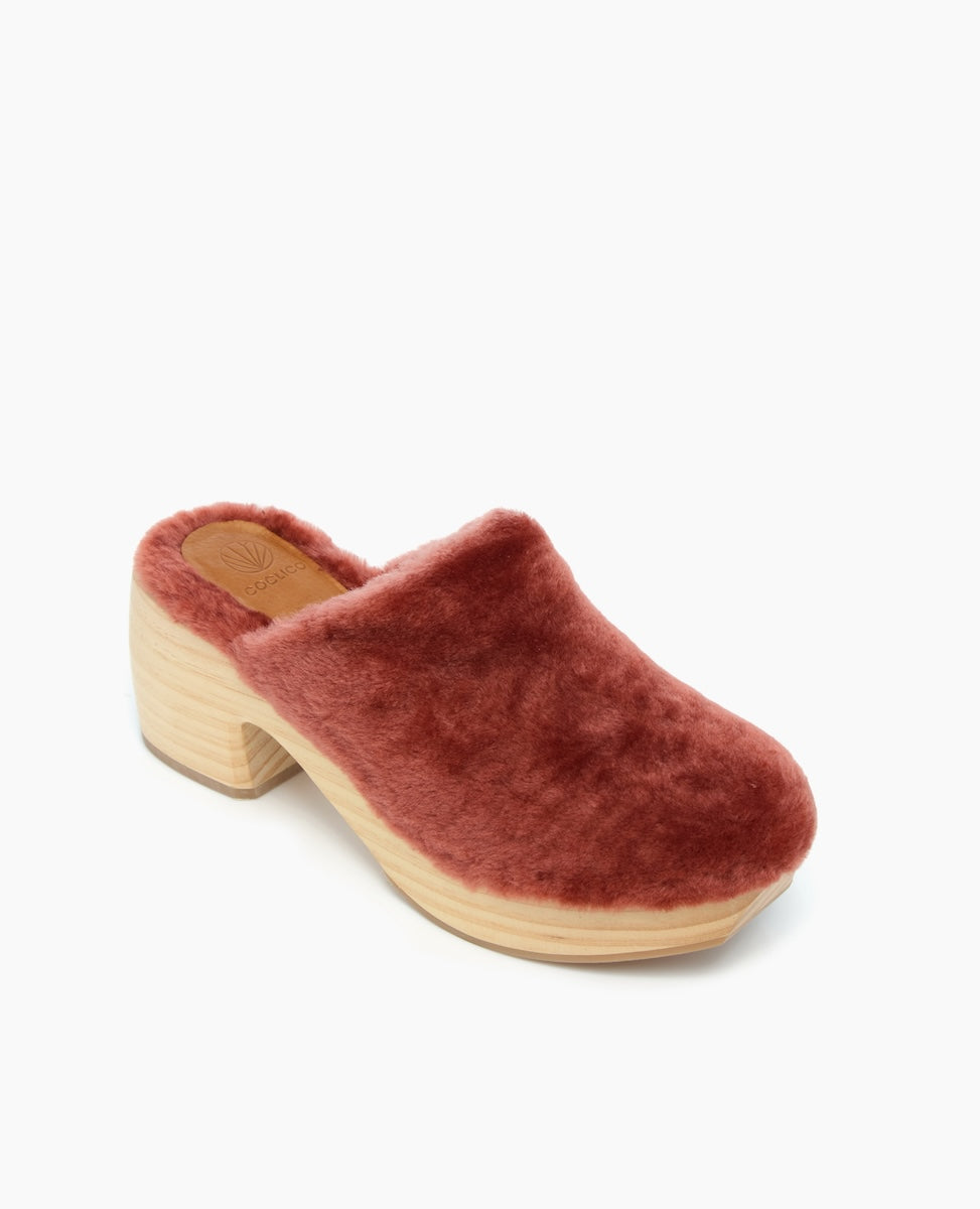 A plush, rust-colored, faux fur clog with a chunky, light wooden heel and platform, featuring a closed toe and an open back.