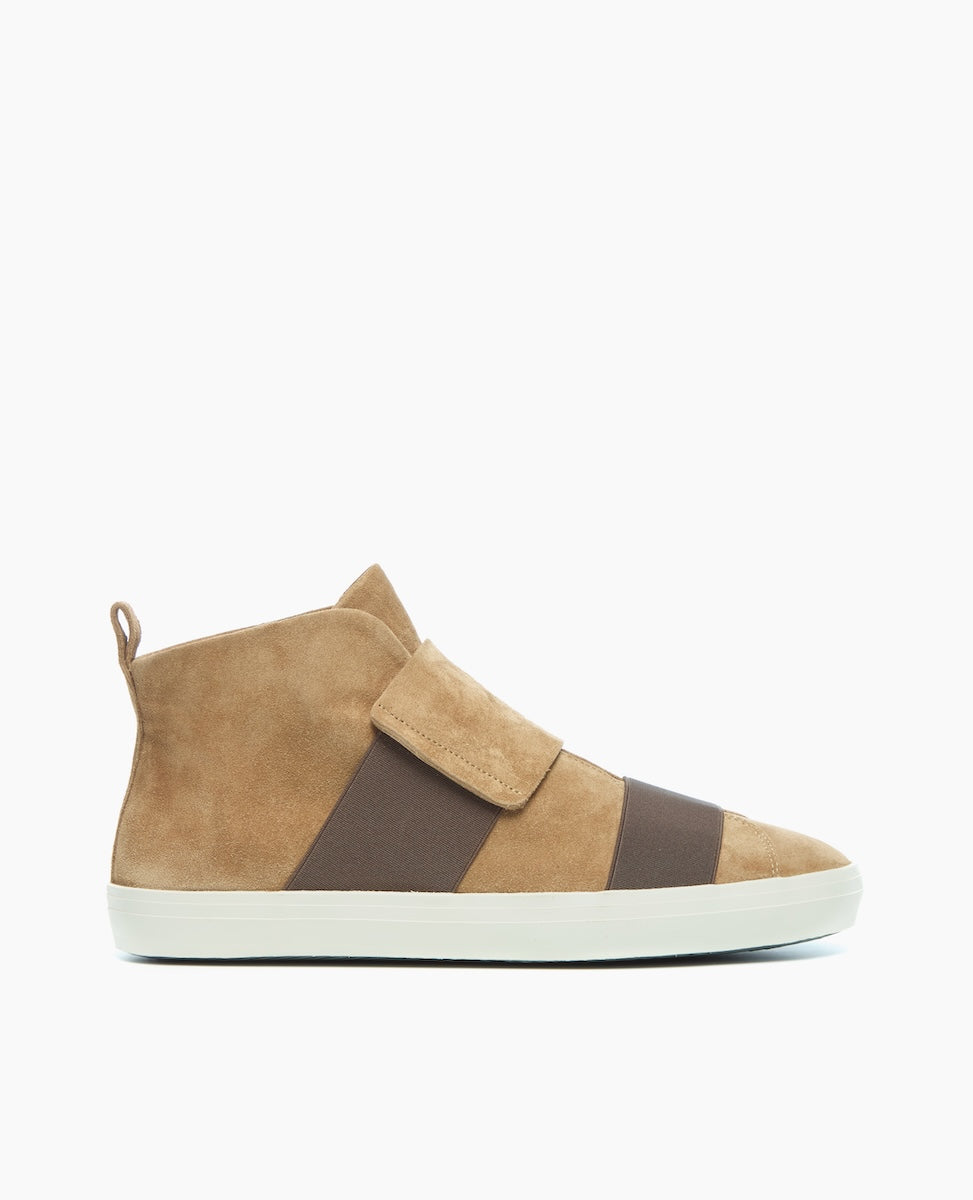 A tan suede high-top sneaker features a wide brown elastic band across the midfoot, a white rubber sole, and a pull tab at the heel.
