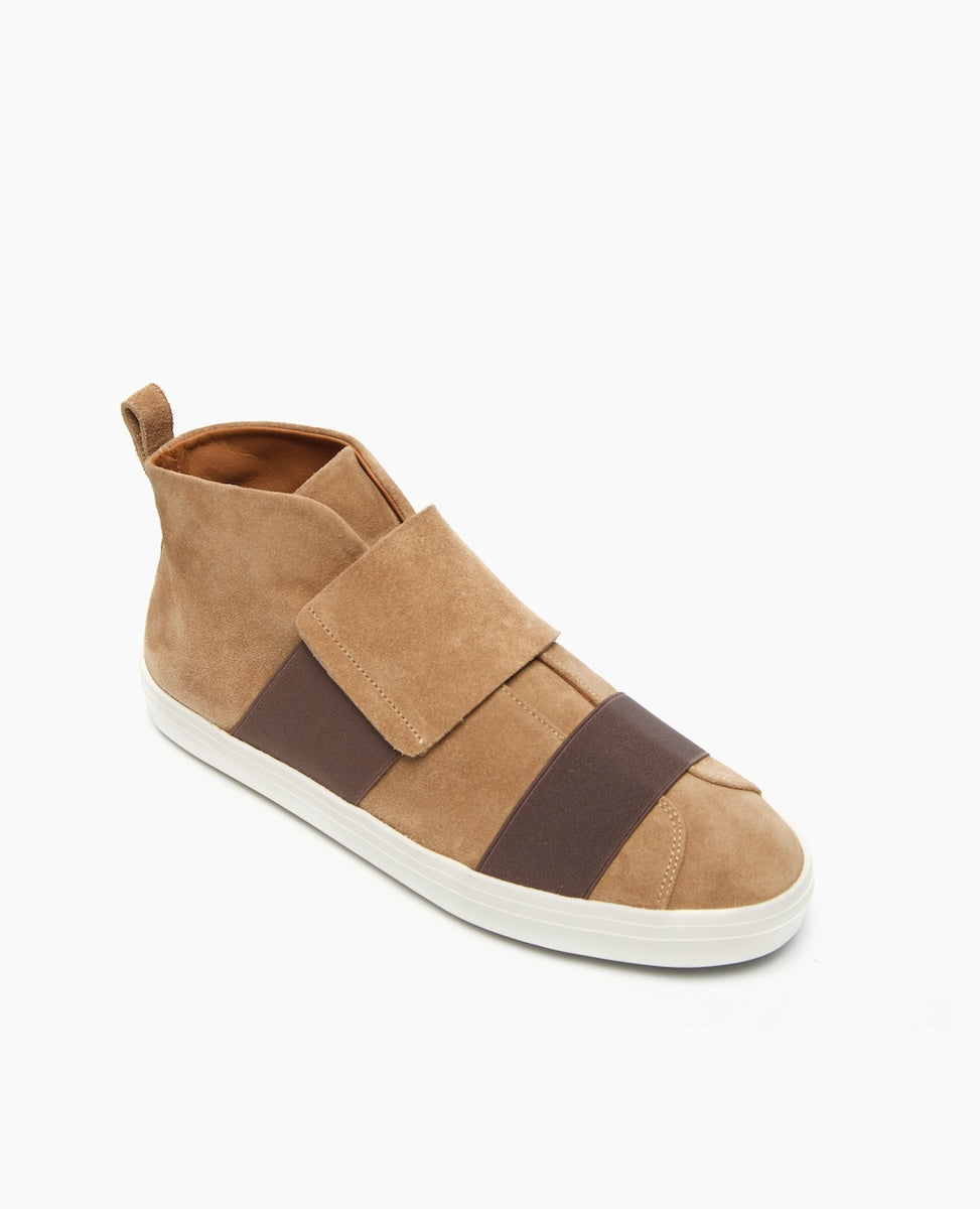 A tan suede high-top sneaker features a sleek design with wide brown elastic bands across the top, a white rubber sole, and a pull tab at the heel for easy wear.