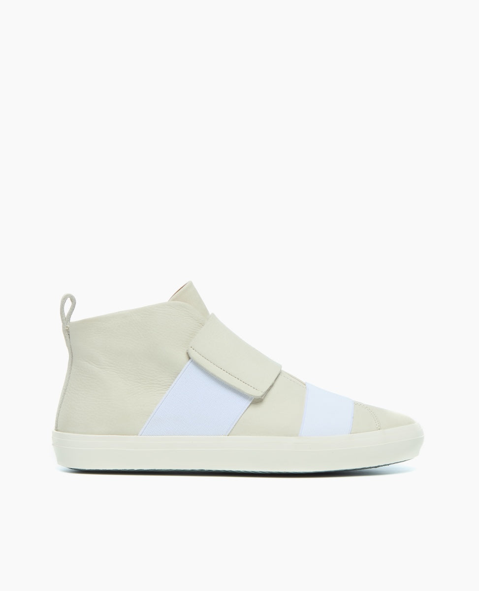 A sleek, high-top sneaker featuring a minimalist design with a light beige and white color scheme, smooth leather texture, a wide velcro strap across the top, and a flat rubber sole.