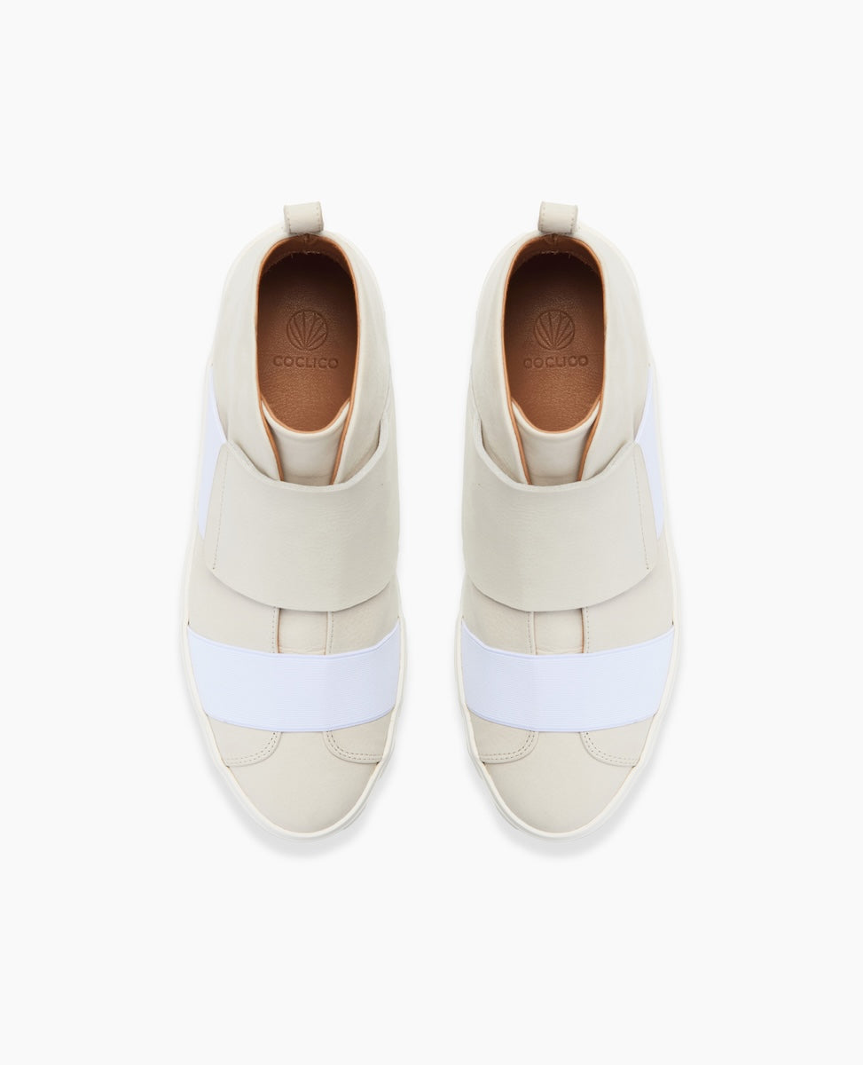 High-top sneakers with a sleek, minimalist design feature a cream and white color scheme, smooth leather material, wide elastic bands across the midfoot, and a subtle logo on the insole.
