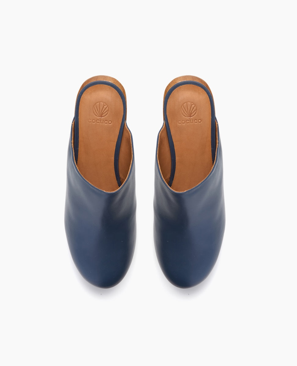 A pair of sleek, navy blue leather mules with a smooth texture, featuring a rounded toe and tan leather insoles embossed with a logo.