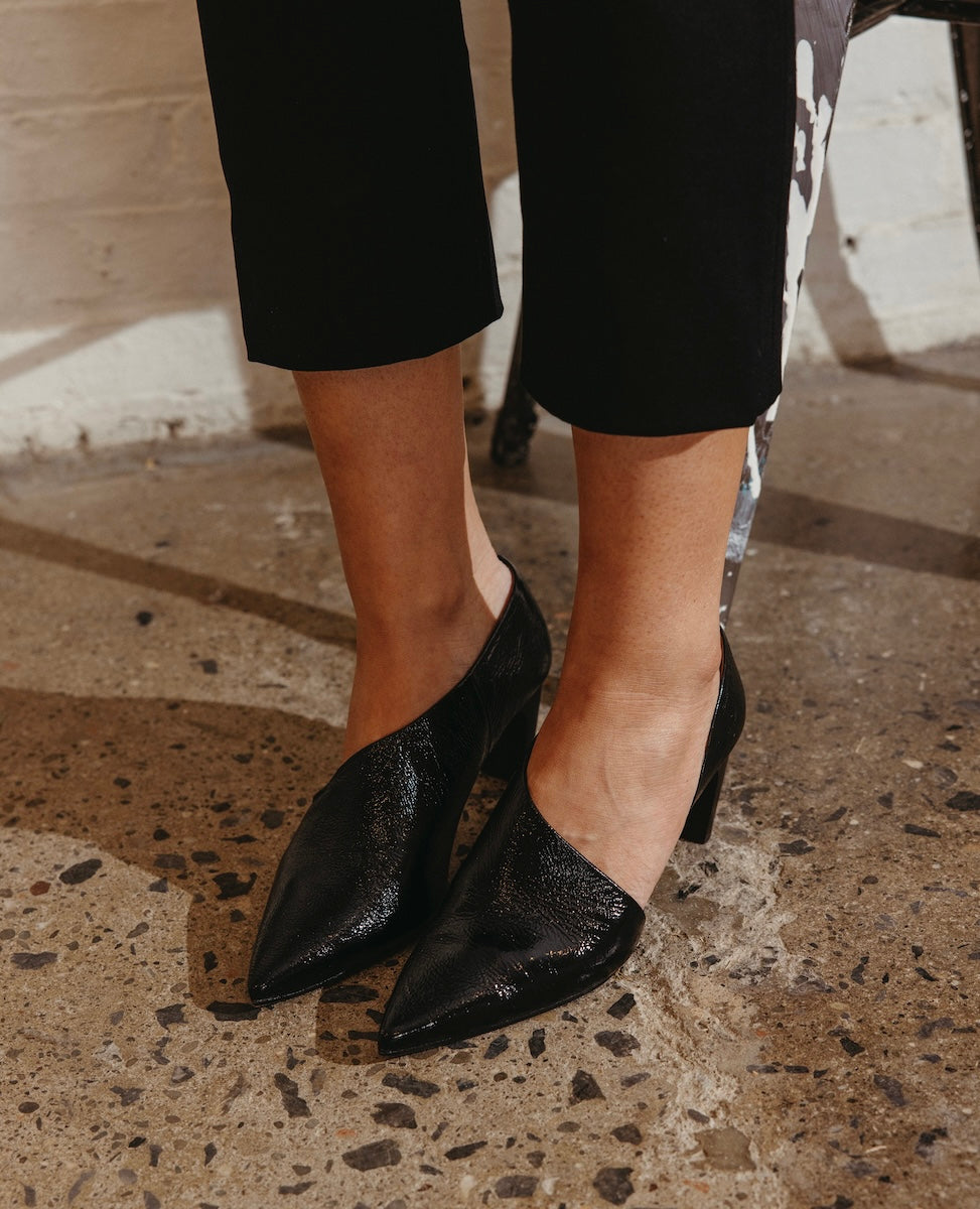Black, pointed-toe high heels with a glossy, textured finish, worn by a person standing on a speckled concrete floor.
