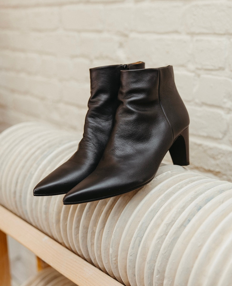 A pair of sleek, black leather ankle boots with pointed toes, a smooth texture, and a moderate heel, displayed on a light-colored, ribbed surface against a brick wall background.