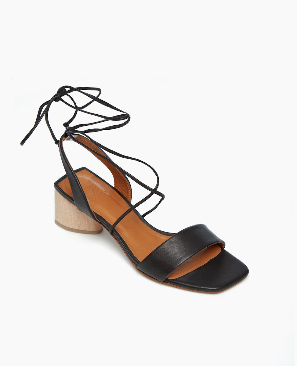 Angled view of the Steph in Black Leather featuring a comfortable leather front band and a winding of leather straps that cascade up the ankle with a tie closure.