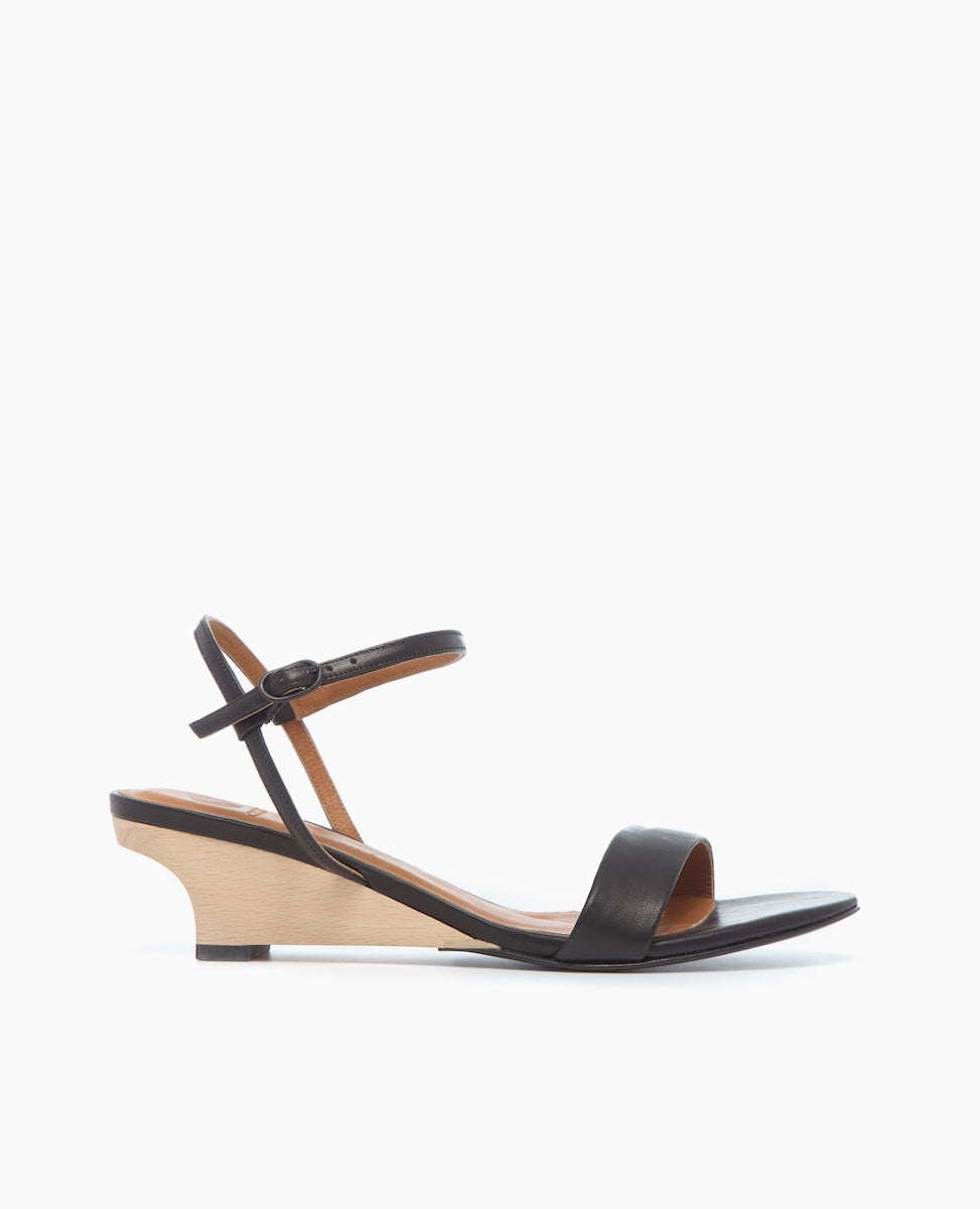 A sleek black sandal with a minimalist design features a thin ankle strap with a buckle, a single toe strap, and a low, light wood wedge heel.