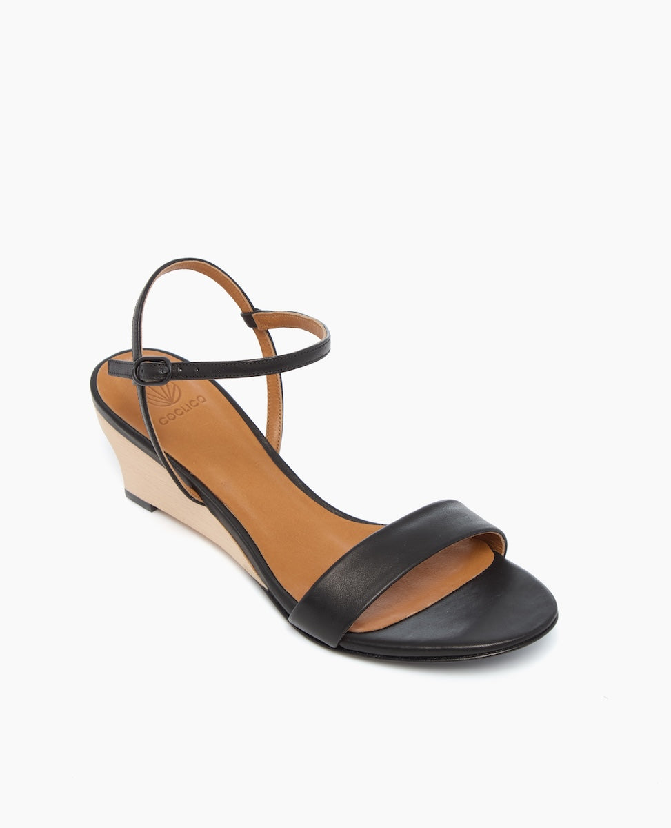 A black leather wedge sandal with a tan insole, featuring a single front strap, an adjustable ankle strap, and a beige wedge heel.