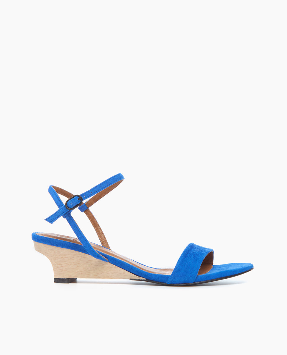 A vibrant blue sandal features a sleek, open-toe design with a thin ankle strap, a low wedge heel in a contrasting light wood texture, and a smooth suede finish.