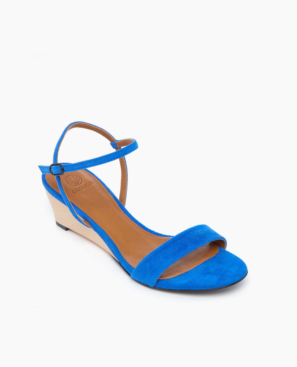 A vibrant blue suede sandal with a low wedge heel, featuring an open toe, a single front strap, and an adjustable ankle strap with a buckle, complemented by a tan insole and a contrasting light beige wedge.