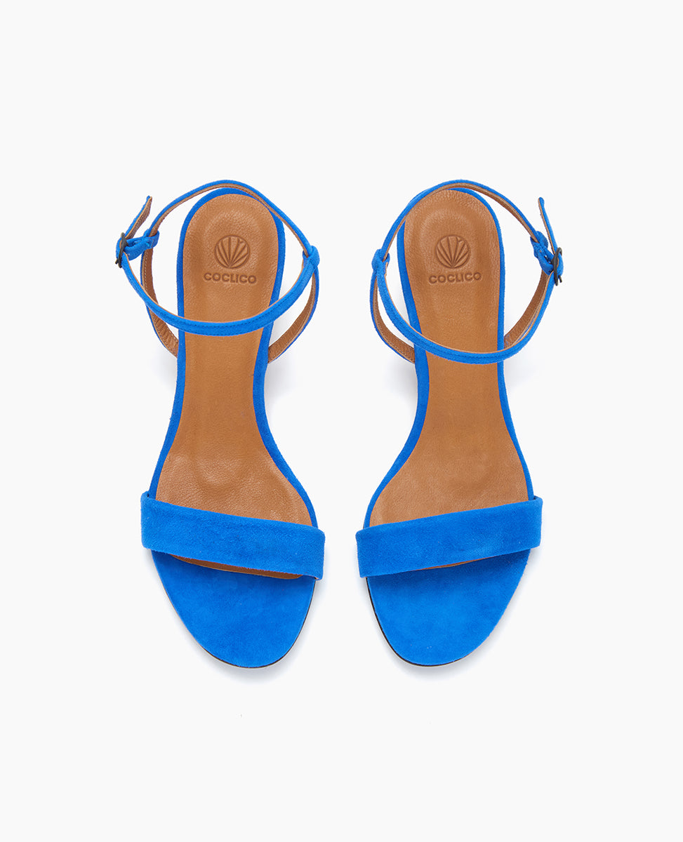 Bright blue sandals with a minimalist design feature a single wide strap across the toes, an adjustable ankle strap with a buckle, and a tan insole with a logo imprint.