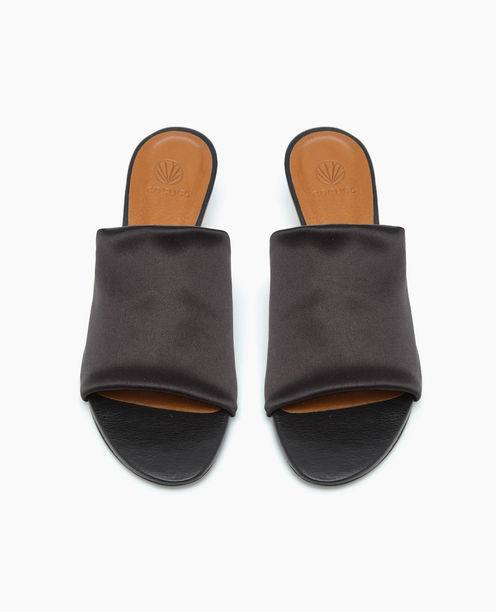 A pair of sleek, black slide sandals with a smooth, wide strap and a tan insole featuring a subtle embossed logo.