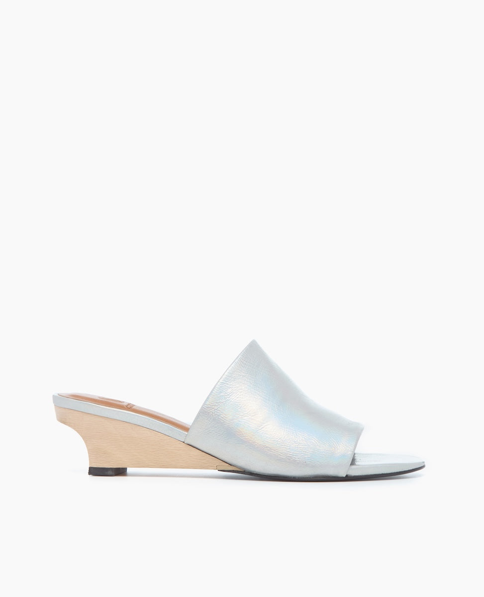 A sleek, open-toe mule sandal featuring a shimmering light blue leather upper, a low wooden wedge heel, and a smooth, minimalist design.