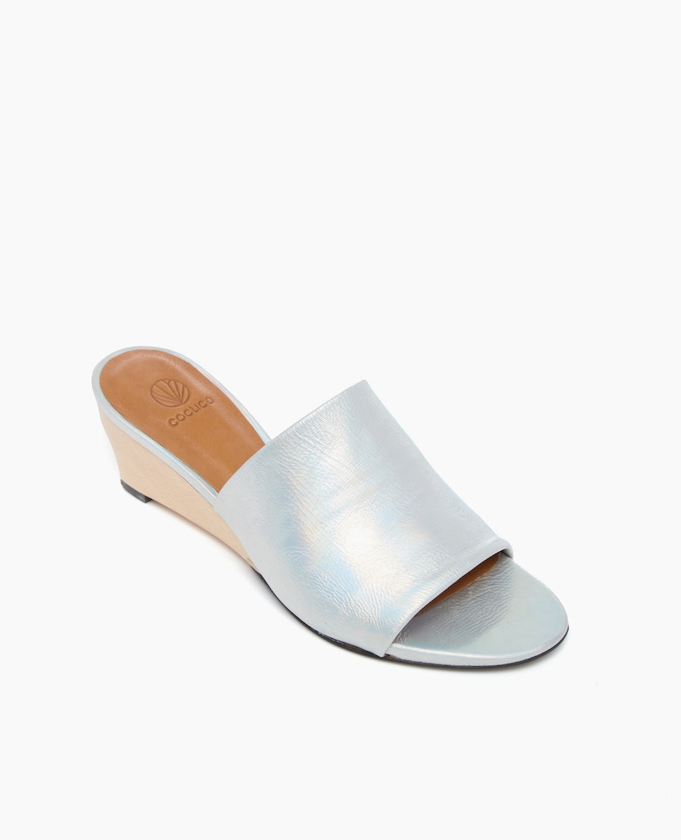 A sleek, open-toe wedge sandal featuring a light blue metallic upper with a subtle textured finish, a tan insole, and a beige wedge heel.