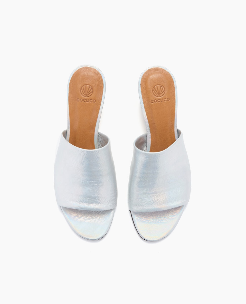 A pair of sleek, open-toed sandals with a metallic silver finish, featuring a smooth leather upper, a tan leather insole with a logo imprint, and a minimalist design with a low heel.