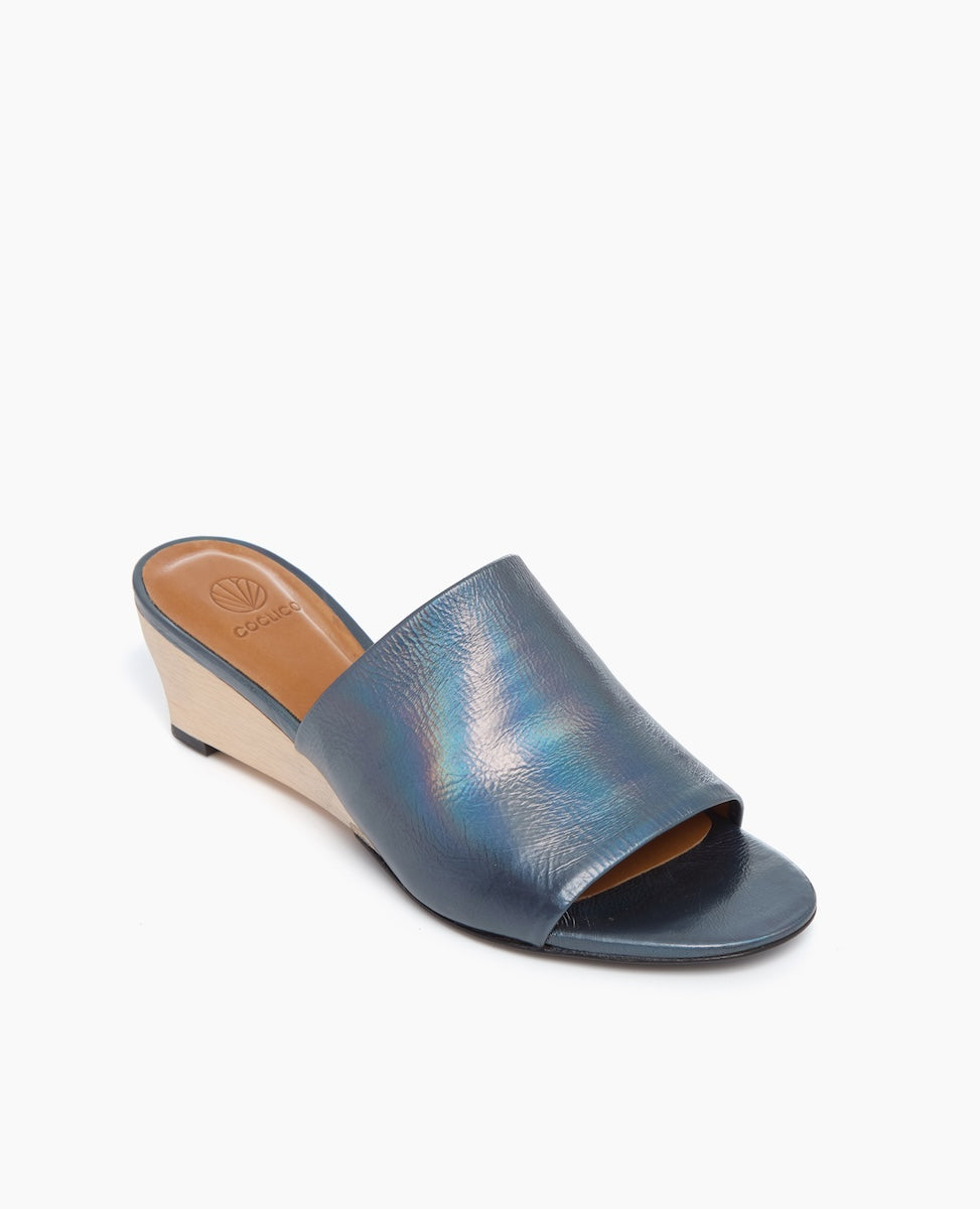 A stylish open-toe wedge sandal featuring a shimmering blue leather upper, a tan insole, and a contrasting light beige wedge heel with a smooth texture.