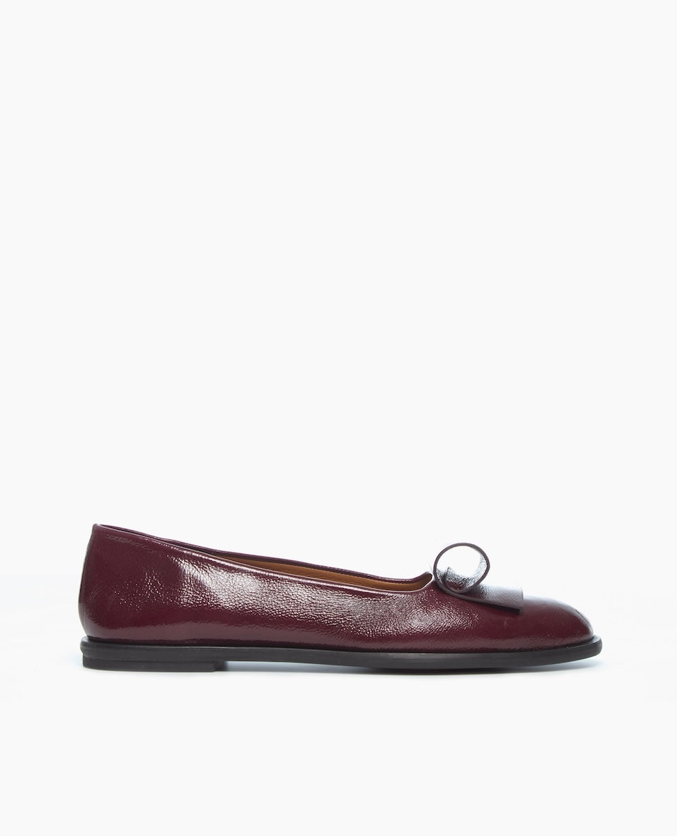 A single burgundy leather flat shoe with an architectural loop detail on the toe, displayed against a plain white background.