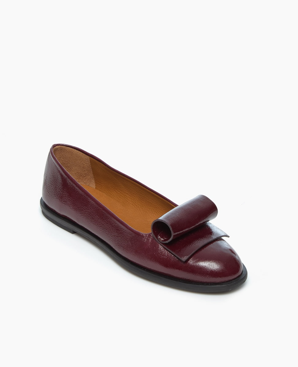 A single burgundy leather flat shoe with a decorative bow on the front and a black sole is displayed against a plain white background.