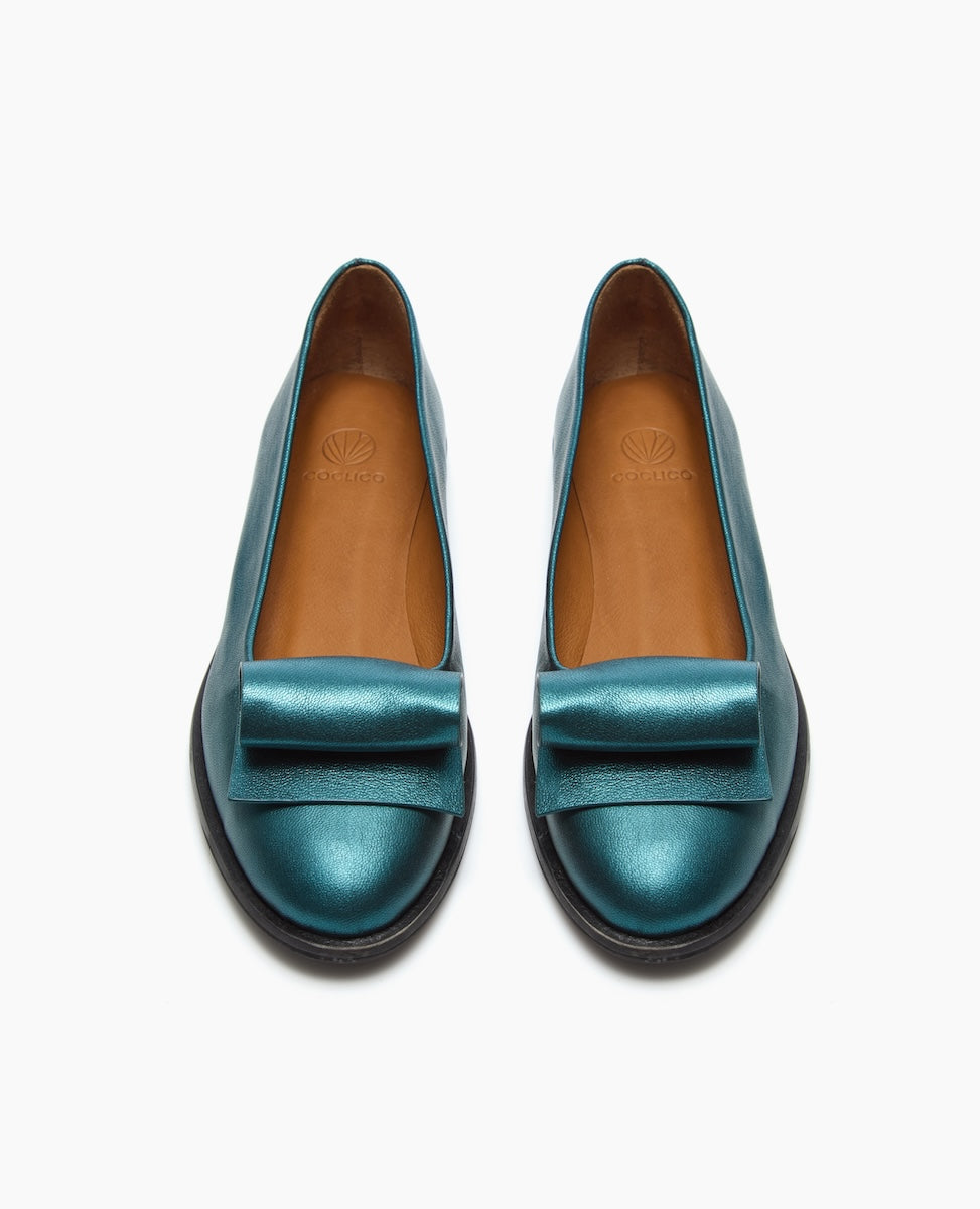 A pair of teal, metallic leather loafers with a smooth texture, featuring an architectural loop detail on the front, a rounded toe, and a black rubber sole.