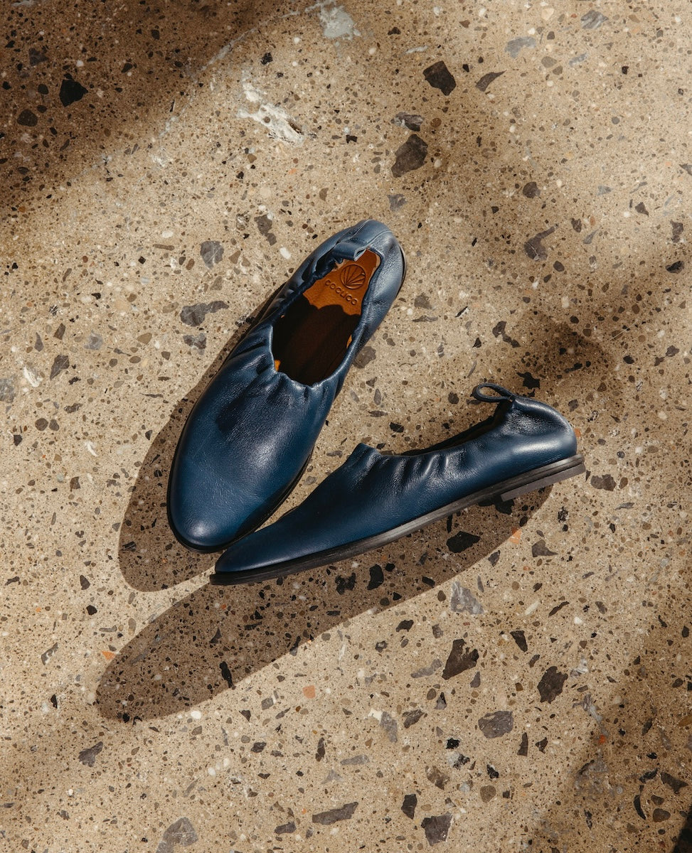 A pair of dark blue, slip-on shoes with elasticized openings are placed on a speckled concrete floor, casting soft shadows.