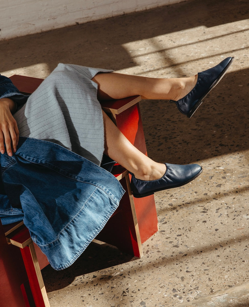 A person wearing a blue dress and black shoes is sitting on a red bench, with sunlight casting shadows on the ground.